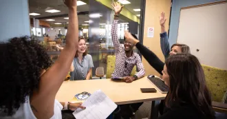 Photo of Madison College students in a meeting room, sitting around a table, raising their hands in excitement.