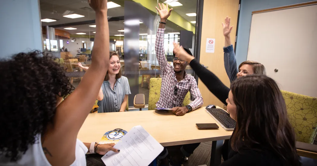Photo of Madison College students in a meeting room, sitting around a table, raising their hands in excitement.