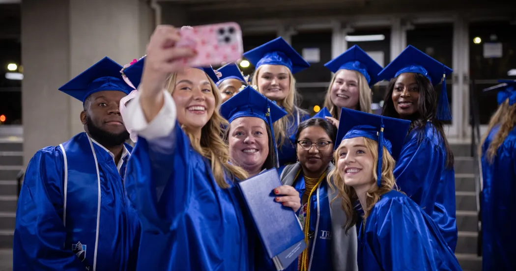 Image of Madison College graduates in blue caps and gown posing for a selfie.