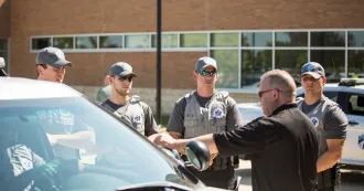 Four criminal justice program students listening to an instructor outside a building and next to a car.