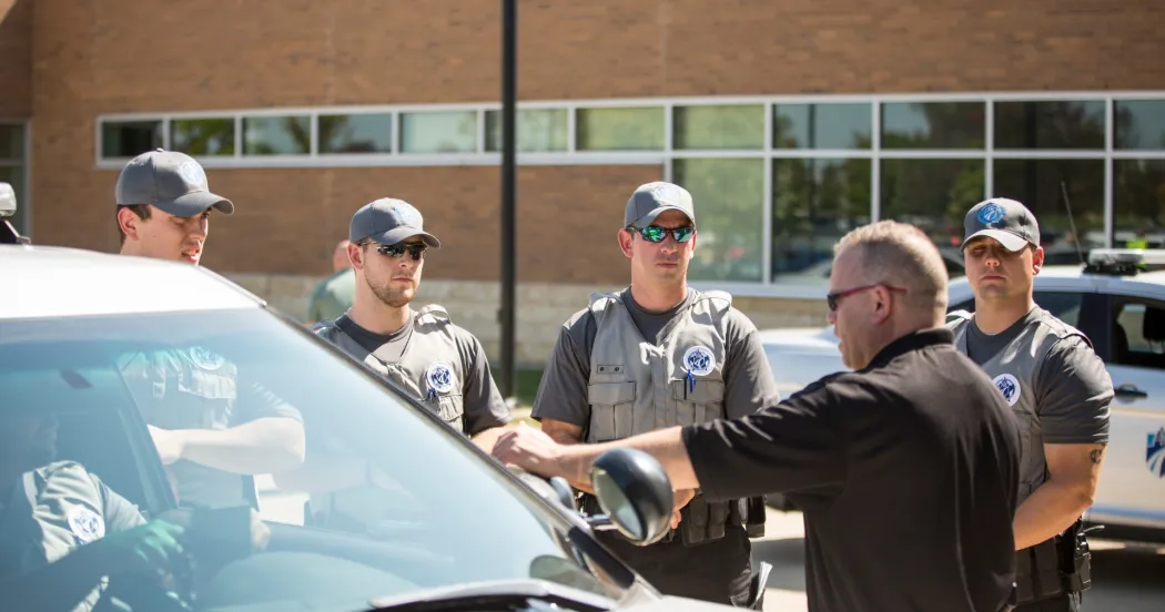Four criminal justice program students listening to an instructor outside a building and next to a car.