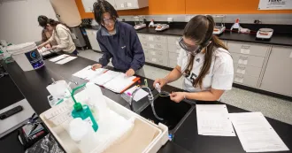 Two students in a lab working on a project, wearing eye googles and putting solution in an eye dropper on a slide.