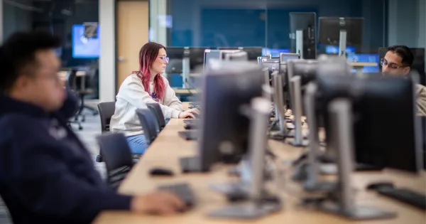 Image of three students, wearing sweatshirts, in front of computer monitors working. 