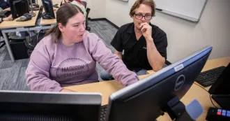 Student, wearing purple sweatshirt, working on desktop computer with instructor by her side.
