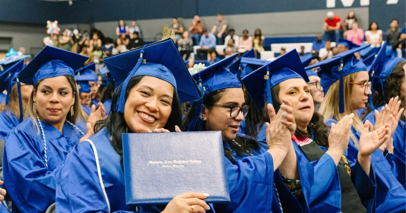 Madison College graduate, wearing a blue cap and gown, holding up her high school equivalency diploma during graduation.