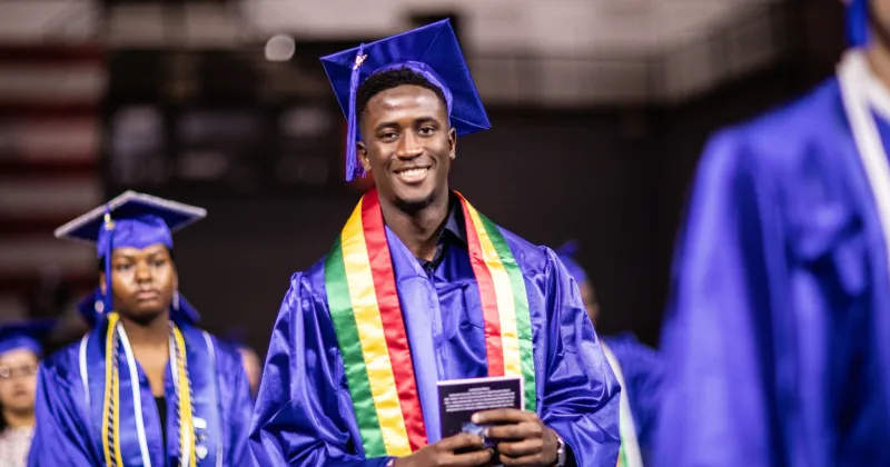 Black graduate,in blue cap and gown and African color shawl, walking in a commencement ceremony.