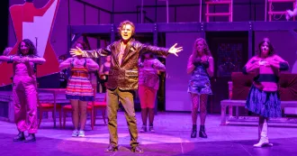 Student in brown coat and pants on a stage singing with arms outstretched.