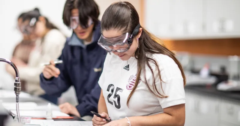 Student in a lab setting wearing protective eye gear and looking at a notebook with a pen in her hand.