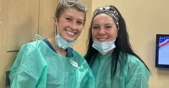 Two dental hygiene students in green scrubs, masks, smiling at camera, in a dental clinic lab.