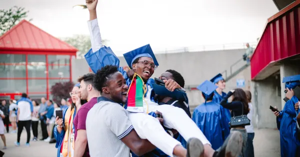 Black male graduate, dressed in blue cap and gown, being held up in the air, by two friends in celebration of his graduation.