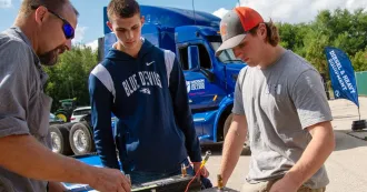 Two male teenagers look on as a instructor demonstrates a battery on a table.