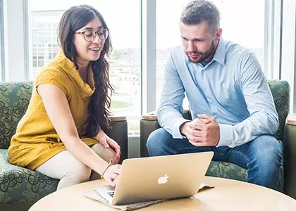 Two students seated at a round table looking at a macbook.